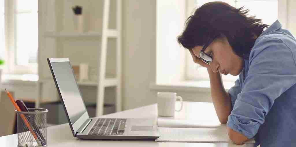 Individual sitting at a desk with a laptop, looking stressed and resting their head on their hand.